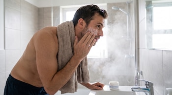 Hombre sin camiseta con toalla en el cuello aplica crema facial de un envase blanco en el lavamanos, con un espejo y una ducha al fondo y vapor en el aire.