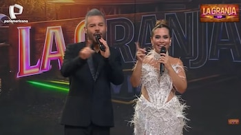 Dos presentadores, un hombre de traje oscuro y una mujer con vestido blanco brillante y plumas, sostienen micrófonos en un escenario con el gran letrero luminoso 'LA GRANJA'