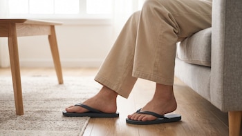 Pies de una persona con sandalias negras y pantalón beige, apoyados sobre un piso de madera junto a un sofá y una mesa baja