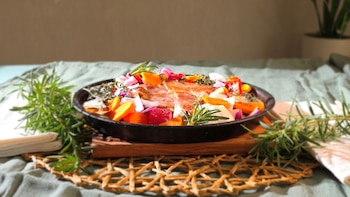 Una sartén negra con trozos de carne, zanahorias, cebolla morada y romero fresco sobre una base de madera y un mantel verde