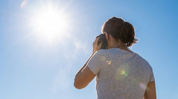 Vista de la espalda de una persona con camiseta gris secándose la cara con una toalla, bajo un cielo azul con el sol brillando intensamente.