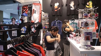 Dos personas en una tienda junto a maniquíes con camisetas impresas, mostradores con llaveros y percheros con ropa.