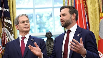 Dos hombres en traje en una sala; el de la derecha, con barba, gesticula al hablar, y el de la izquierda lo mira. Al fondo, banderas y una estatua