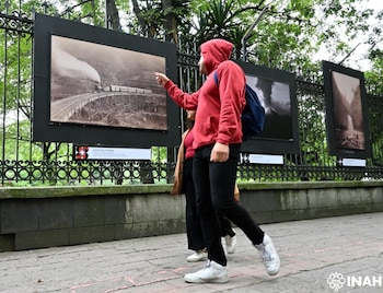 Dos personas observan fotografías enmarcadas colgadas de una reja de hierro. La imagen principal muestra un tren a vapor en un puente de madera, al aire libre