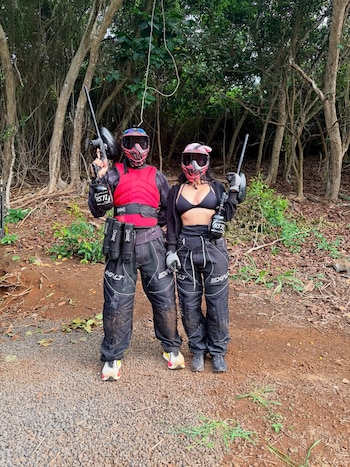 Dos personas, máscaras, chalecos, pantalones, armas de paintball, terreno con tierra y grava, árboles y vegetación
