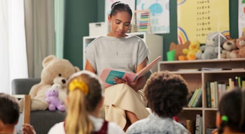 Mujer lee un libro a niños pequeños sentados frente a ella. El fondo muestra una estantería con libros y peluches, y carteles educativos