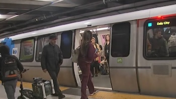 Vista de un tren de tránsito rápido estacionado en la plataforma de una estación, con personas subiendo y bajando; el tren muestra el destino 'Daly City'