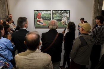 Vista trasera de un grupo de personas en una galería de arte, mirando dos cuadros grandes en una pared blanca que muestran figuras en un paisaje verde. Una mujer sostiene un micrófono