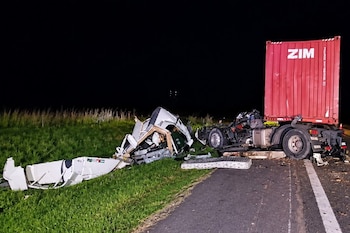 Restos de un camión blanco y un contenedor rojo con logo ZIM en una carretera con pasto verde oscuro de noche. Hay escombros dispersos