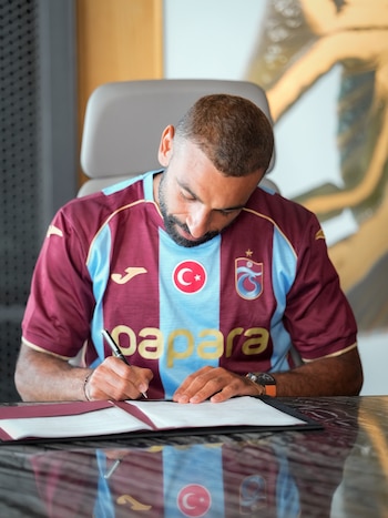Un hombre con barba y camiseta de fútbol azul y granate con los logos de Turquía y Trabzonspor firma un documento con un bolígrafo sobre una mesa