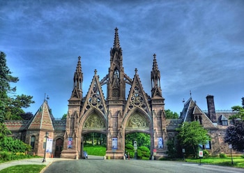 Gran arco de entrada de piedra de estilo gótico del Cementerio Green-Wood con múltiples pináculos y relieves escultóricos bajo un cielo azul claro, flanqueado por árboles
