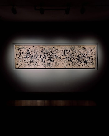 Pintura rectangular de arte abstracto con salpicaduras negras y rojas sobre un fondo crema claro, enmarcada y colgada en una pared oscura bajo focos de techo