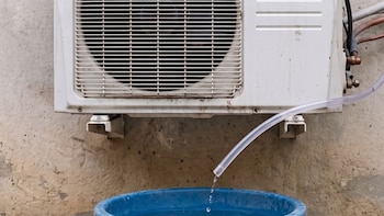 Unidad exterior de aire acondicionado blanca con rejilla y ventilador, manguera transparente descargando gotas de agua en un cubo azul, sobre una pared beige