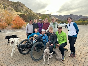 Grupo de diez personas y seis perros posa con una bicicleta reclinada. Fondo de montañas, árboles con follaje otoñal y una camioneta blanca