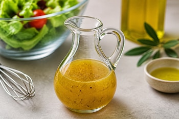 Una jarra de vidrio con vinagreta, un batidor de varillas, un cuenco con ensalada y una botella de aceite de oliva.