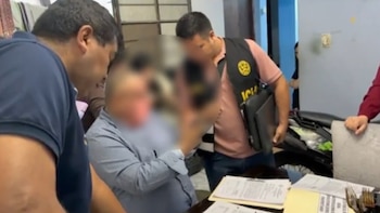 Varias personas y agentes del orden rodean a un hombre con el rostro pixelado sentado frente a una mesa llena de documentos en un espacio interior