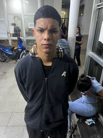 Un hombre joven, Lautaro Nicolás Damián Brest, de pie, mirando fijamente a la cámara, con una sudadera oscura. Al fondo, personas y una motocicleta azul en un interior