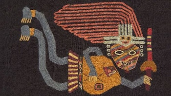 Primer plano de un textil Paracas con un bordado multicolor de una figura antropomórfica. Presenta una cabeza con tocado, cuerpo naranja y extremidades grises sobre fondo oscuro