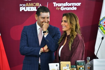 Un hombre con traje azul y una mujer con camisa guinda conversan detrás de una mesa con frascos y paquetes de alimentos
