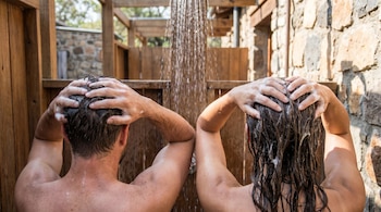 Un hombre y una mujer, vistos de espaldas, se lavan el cabello con champú bajo dos duchas. Se ven sus cabezas, brazos y la espuma en su pelo. Gotas de agua caen.