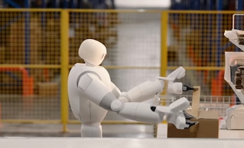 Robot humanoide blanco que sostiene una caja de cartón sobre una mesa en un almacén con rejas amarillas