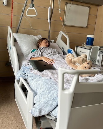 Joven paciente durmiendo en cama de hospital blanca con sábanas estampadas. Un suero intravenoso, una laptop y un oso de peluche están junto a ella