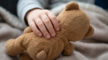 Primer plano de la mano de un niño de unos tres años tocando un osito de peluche marrón sin adornos sobre una manta beige.