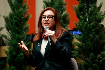 Mujer de cabello rojizo, gafas y traje oscuro hablando en un micrófono. Detrás, dos árboles verdes tipo pino y una pared roja