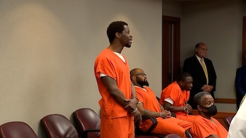 Un hombre afroamericano con uniforme naranja de prisión está de pie. Detrás de él, otros hombres sentados también visten uniformes naranjas. Dos hombres con traje observan en el fondo