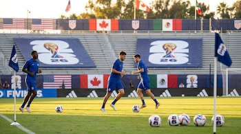 Tres futbolistas en uniforme azul calientan en un campo de fútbol con balones oficiales del Mundial 2026. Banderas de EE.UU., Canadá y México en el estadio.