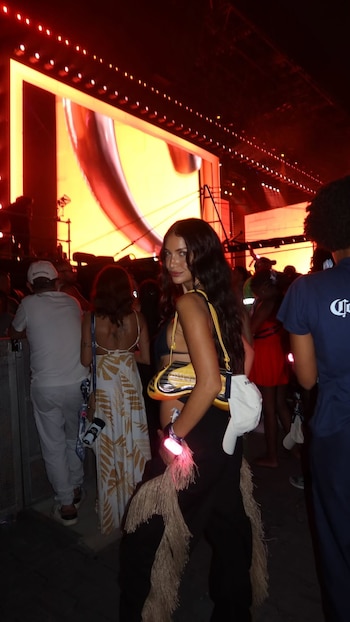 Zaira Nara, de perfil, mirando a la cámara, en un concierto con luces rojas y una pantalla gigante de fondo. Lleva un top negro, pantalones con flecos y un bolso amarillo