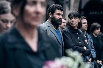 Un hombre con barba y una mujer con chaqueta oscura miran hacia el frente junto a otras personas en la calle