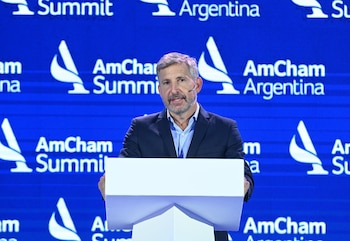 Hombre de mediana edad con cabello gris y barba, vestido con traje, habla en un podio blanco frente a una pantalla LED azul con el logo de AmCham Argentina