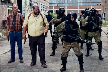 Dos hombres con pistolas, uno con camisa a cuadros y otro con camisa amarilla. Un grupo de personas con uniformes de camuflaje, cascos, pasamontañas y rifles. Un casco lleva una GoPro. Fondo con edificios de ladrillo
