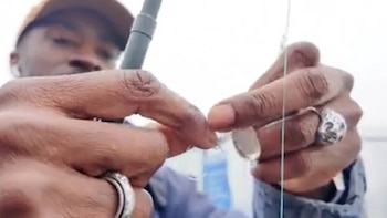 Primer plano de las manos de un hombre con piel oscura y varios anillos, manipulando un hilo fino. El rostro del hombre y parte de una caña de pescar se ven borrosos al fondo