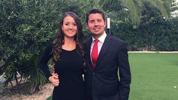 Un hombre con traje oscuro y corbata roja sonríe junto a una mujer con vestido negro y cabello largo frente a un fondo de vegetación verde y césped