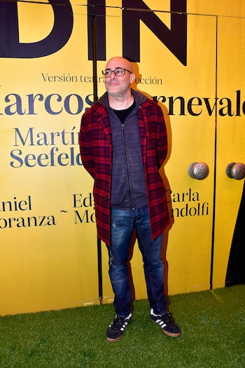 Hombre calvo con gafas, chaqueta a cuadros roja y azul, jeans y zapatillas deportivas, de pie frente a un fondo amarillo con letras negras grandes y césped artificial