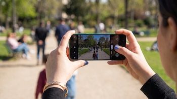 Manos sujetando un teléfono inteligente verde que muestra en pantalla la vista de la cámara enfocada hacia un camino en un parque.