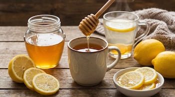 Una jarra de miel y un dosificador goteando miel en una taza. Limones enteros y en rodajas. Un vaso de agua caliente con limón sobre una mesa de madera.