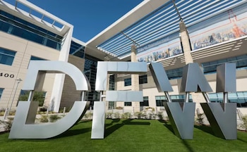 La gran señal metálica con las letras DFW se erige sobre césped verde frente a un edificio moderno de cristal y hormigón bajo un cielo azul claro