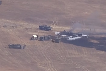 Vista aérea de un terreno desértico con vehículos de emergencia y figuras humanas alrededor de un área humeante con espuma