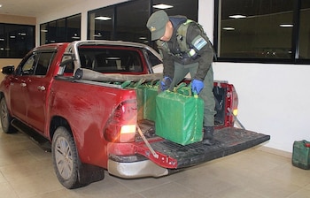 Oficial uniformado con guantes azules saca paquetes rectangulares verdes de la caja de una camioneta pickup roja, con la compuerta trasera abierta