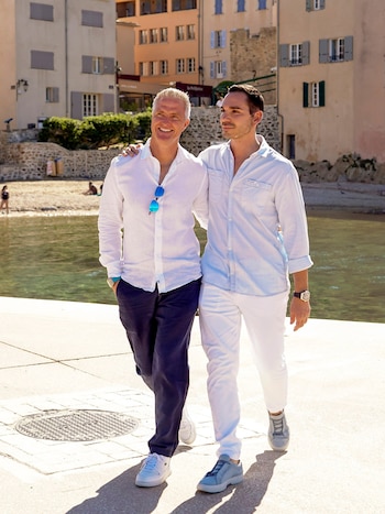Ralf Schumacher, de cabello blanco, y un hombre joven, de cabello oscuro, caminan juntos sonriendo por un paseo marítimo bajo el sol, con el mar y edificios de fondo