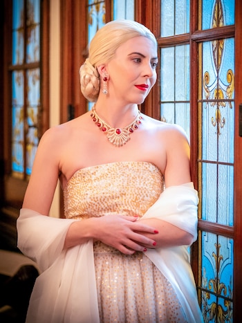 Mujer rubia de perfil con vestido de lentejuelas doradas, collar de rubíes y diamantes, aretes, y chal blanco, frente a un vitral ornamental