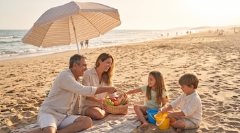 Dos adultos y dos niños sentados en la arena de una playa junto a una cesta de comida, cubos de plástico y una sombrilla.