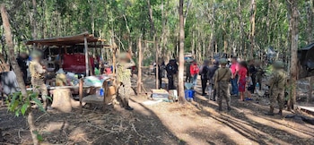 Grupo de personas uniformadas y civiles en área boscosa; incluye árboles, vegetación, estructuras de madera y lona, bolsas, bidones y un vehículo