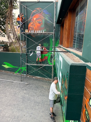 Tres personas pintan con aerosol un mural con un rostro femenino y mariposas sobre una pared exterior junto a un andamio metálico