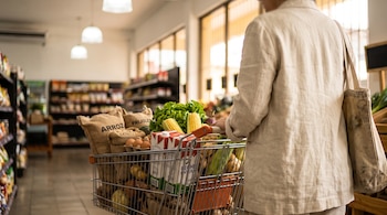 Persona de espaldas empujando un carrito de compras con sacos de arroz, frijoles, leche, huevos y verduras en un pasillo.