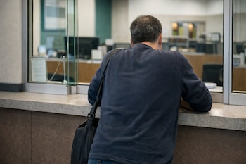 Vista trasera de un hombre adulto con camiseta azul oscuro y bolso al hombro, apoyado en un mostrador. Una partición de vidrio lo separa de un área de oficina.