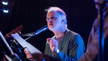 Hombre de cabello gris y barba, vestido con cárdigan verde, frente a un micrófono, sostiene papeles en un escenario con iluminación azul y una guitarra al fondo